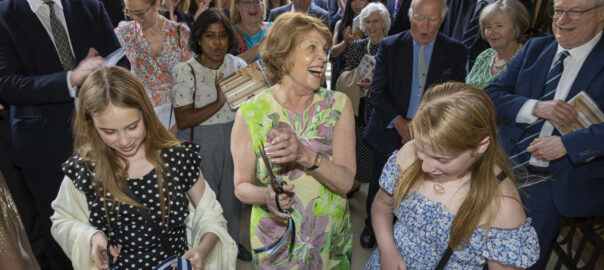 Dagmar Dolby, standing in front of a group of Fellows and alumni, and between her two granddaughters, laughs after cutting the ribbon to open Phase 1 of the Mill Lane site